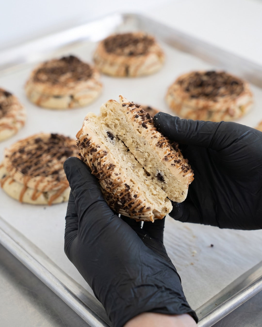 Chilled Biscoff Oreo cookie broken in half in black-gloved hands over a bakery sheet tray